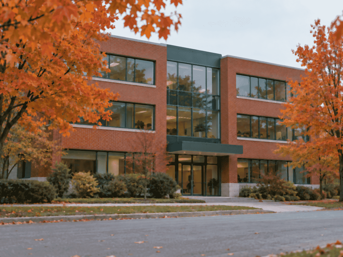 Un immeuble de bureaux de trois étages en brique rouge avec une entrée vitrée, des érables orangés devant, en automne, sans enseigne