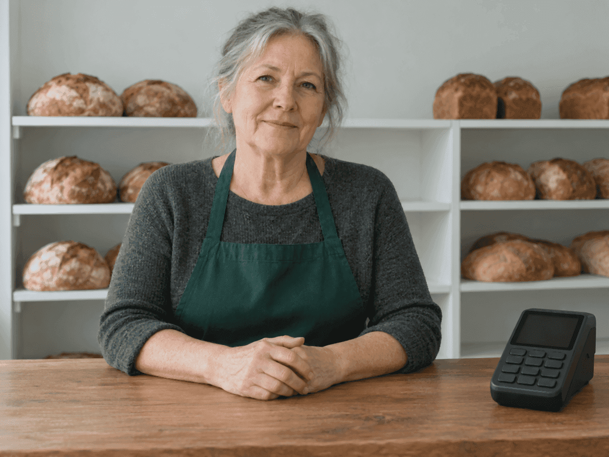 La propriétaire d’une petite boulangerie, dans la soixantaine, en tablier vert derrière un comptoir en bois, des miches sur les tablettes derrière elle et un terminal de paiement près de ses mains
