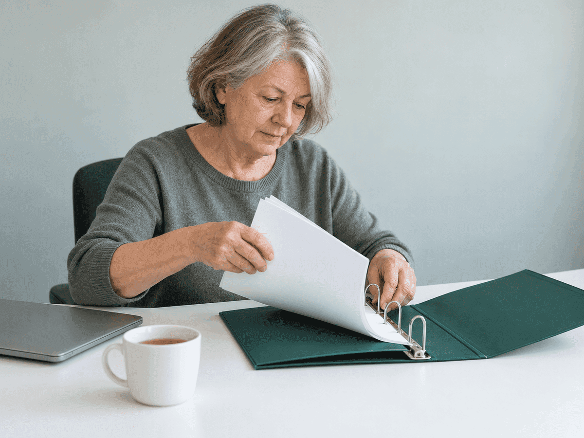 Une administratrice dans la soixantaine ajoute des feuilles blanches dans un classeur à anneaux vert ouvert, sur un bureau blanc, un portable fermé et une tasse de thé à côté d’elle