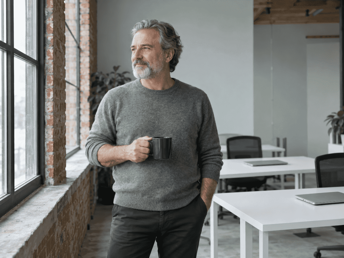Le fondateur d’une petite entreprise de logiciels, dans la fin cinquantaine, debout à la fenêtre d’un bureau-loft en brique, une tasse de café à la main, des bureaux libres avec des portables fermés derrière lui