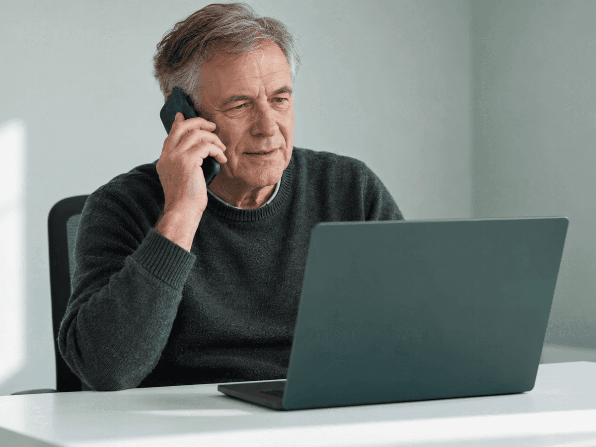 A business owner in his sixties on his mobile phone at a white desk, his open laptop facing him and out of view