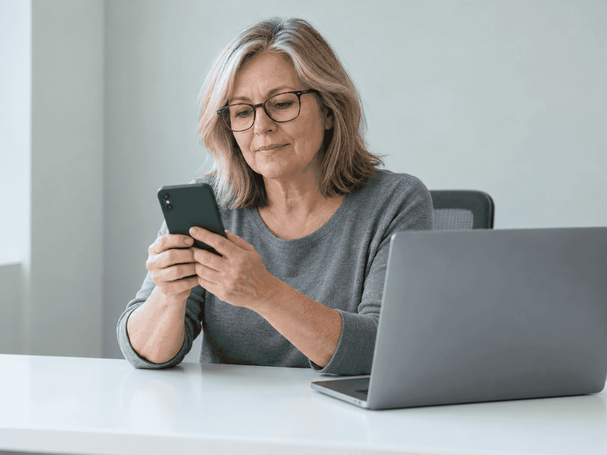 Une gestionnaire de bureau dans la fin cinquantaine, en lunettes, regarde son téléphone à un bureau blanc, son portable ouvert tourné dos à l’objectif