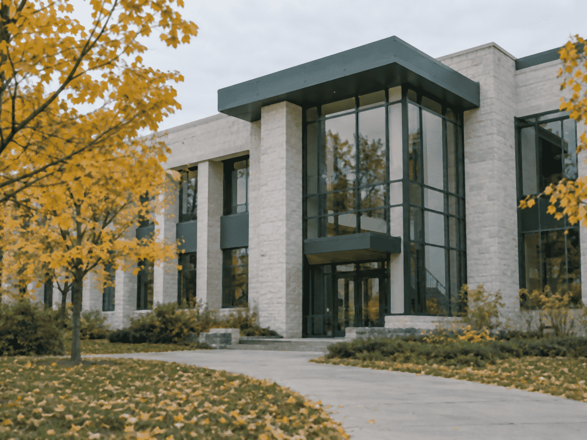 A pale stone college building with a tall glass entrance, yellow autumn trees and fallen leaves on the lawn, no signs or logos