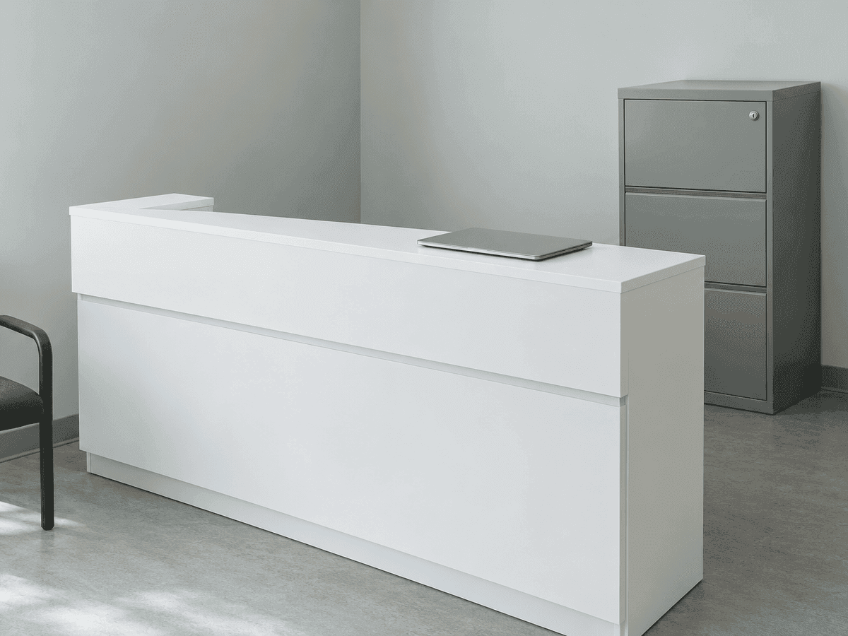 Un comptoir d’accueil blanc inoccupé avec un portable fermé, un classeur gris à serrure derrière et une chaise d’attente sur le côté